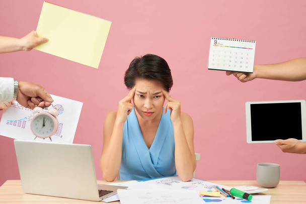 Overworked young employee refuses all things, frowns face in annoyance, sits at desktop with paper documents and notepad, isolated over pink background. Female workes bothered by many questions - Foto, Imagen