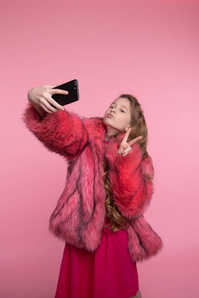 Beautiful girl takes a selfie and shows a gesture of peace in fashionable clothes in a fur coat in pink fur - Photo, image