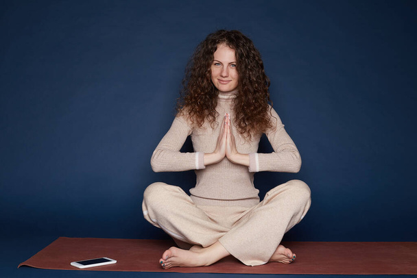Спокойная женщина с мохнатой прической, одетая в спортивную одежду, медитирует на полу в пустой комнате, слушает уроки духовной практики на мобильном устройстве, позирует в позе лотоса, пытается расслабиться. Концепция йоги
. - Фото, изображение