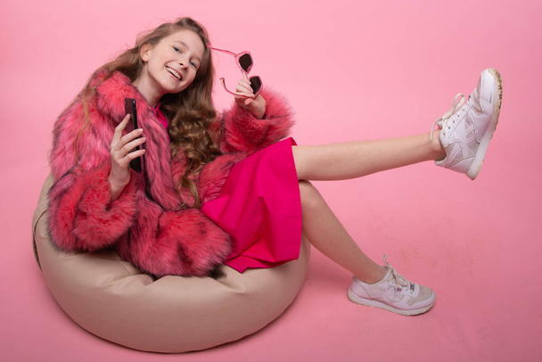 Beautiful young girl sitting in an armchair beige bag listens to music on the phone. Fashionable pink fur coat, pink dress - Foto, afbeelding