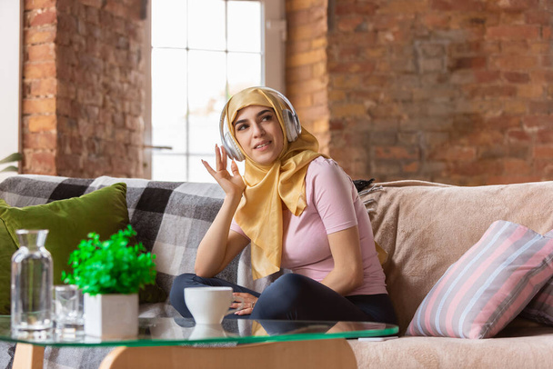 Μια όμορφη νεαρή μουσουλμάνα στο σπίτι κατά τη διάρκεια της καραντίνας και της αυτο-μόνωσης, χρησιμοποιώντας ακουστικά, να ακούσετε μουσική, απολαμβάνοντας - Φωτογραφία, εικόνα
