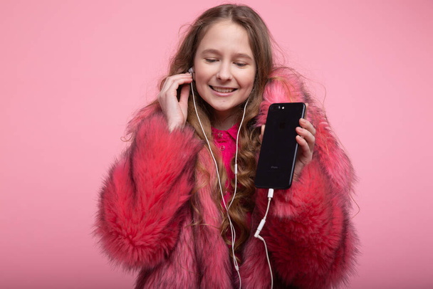 Beautiful young girl inserts headphones into ears in a pink coat on a pink background. - Zdjęcie, obraz