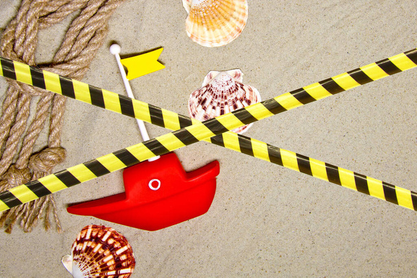 Taśma ochronna - kwarantanna, izolacja, zakaz wstępu. Seashells i dekoracji liny morskiej na tle piasku z czerwonym statku zabawki. - Zdjęcie, obraz