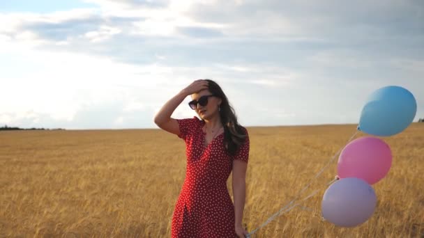 小麦畑を背景にサングラスをかけ、風船を手にした若い女性。赤いドレスの美しい女の子は大麦のプランテーションで彼女の長い茶色の髪をまっすぐにします。スローモーション - 映像、動画