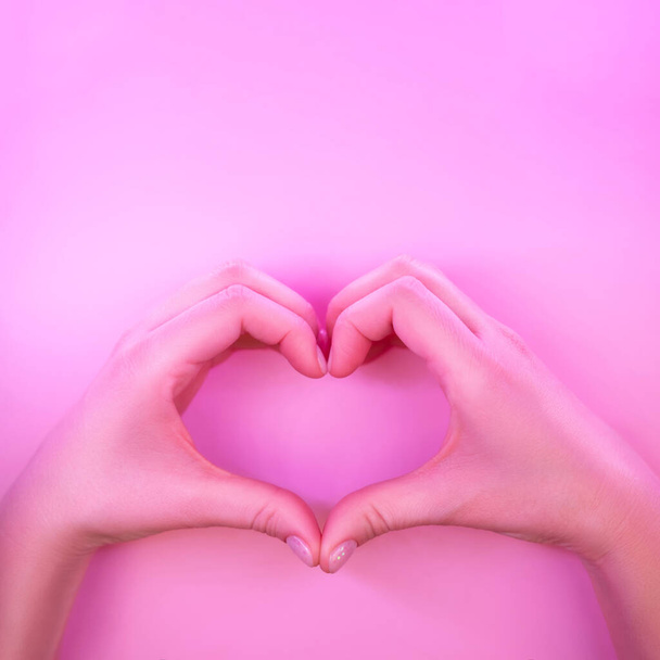 Young woman's hands made gesture of heart shape in trendy neon light on pink background. Love concept. - Foto, afbeelding