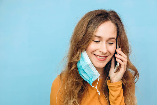 Ένα όμορφο κορίτσι στο τηλέφωνο με ιατρική μάσκα. Πορτραίτο ενός κοντινού πλάνου ενός κοριτσιού σε μπλε φόντο. Κόβιντ-2019. Πανδημία 2020. Το κορίτσι επικοινωνεί συναισθηματικά στο τηλέφωνο.. - Φωτογραφία, εικόνα