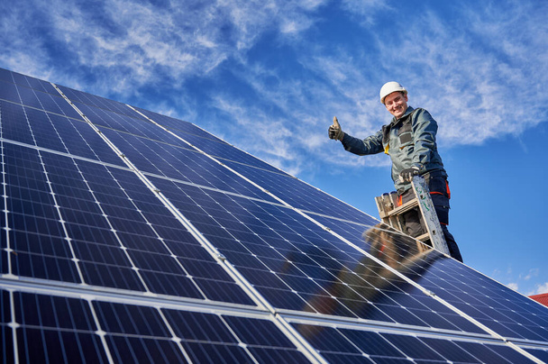 Portrait en angle bas d'un travailleur souriant, installant des piles solaires, debout sur une échelle à la centrale solaire contre le ciel bleu, montrant le pouce levé. Concept de sources d'énergie alternatives
. - Photo, image