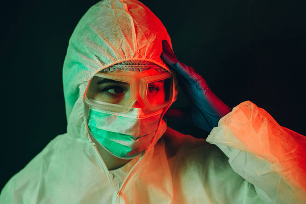 young girl doctor in protective clothing on a black background. Gloomy lighting. Tired doctor. Epidemic covid-2019. Portrait of a doctor in glasses, gloves and a mask. stop the spread of coronavirus. - Photo, Image