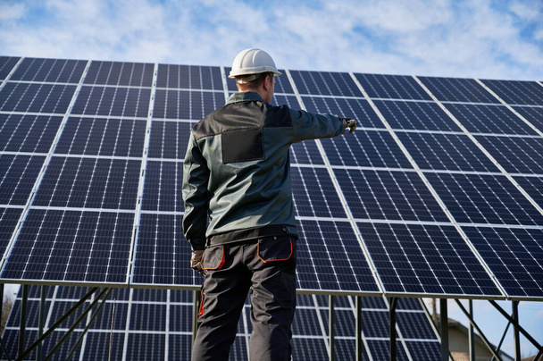 Low angle view on technician in dark blue uniform standing with his back to the camera, pointing at solar plant. Rear view. Alternative ecological source of energy concept - Foto, imagen