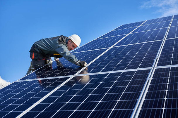 Beautiful and shiny solar battery surface and man worker wearing a uniform, standing on ladder. Male electrician installing solar modules on sunny day, low angle view. Concept of alternative energy - Photo, Image