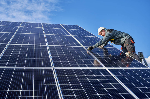 Migawka pozioma pięknej i błyszczącej powierzchni baterii słonecznej oraz męski pracownik noszący mundur, stojący na drabinie, instalujący moduły słoneczne w słoneczny dzień, widok z niskiego kąta - Zdjęcie, obraz