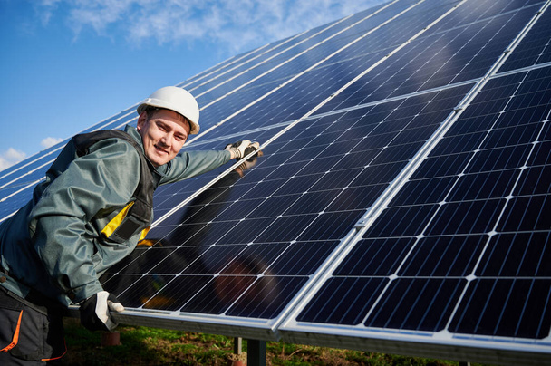 Homem alegre eletricista no capacete de segurança reparando módulo solar fotovoltaico. Trabalhador masculino olhando para a câmera e sorrindo, mantendo o sistema de painel solar fotovoltaico. Conceito de energia alternativa
 - Foto, Imagem