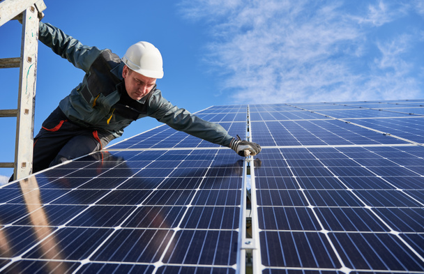 Man electrician standing on ladder and installing photovoltaic solar panels under blue cloudy sky. Technician wearing safety helmet and gloves. Concept of alternative energy and power - Photo, Image