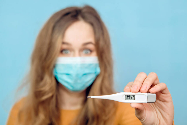 Close-up portrait of a girl in a medical mask on her face with suspected coronavirus and arm with outstretched thermometer. 2020 coronavirus epidemic. Covid-2019. Woman on a blue background. - Foto, Imagen