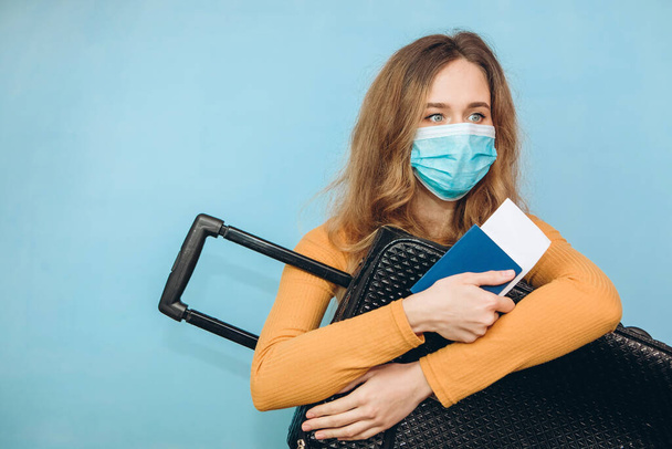 Menina turista em uma máscara médica senta-se com bagagem em suas mãos em um fundo azul. Surto de coronavírus no COVID-19. Viagens e conceito de coroa. Um turista não pode sair devido a uma pandemia
. - Foto, Imagem