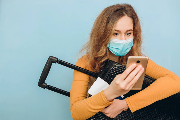 Девушка-турист в медицинской маске сидит с багажом в руках на синем фоне. COVID-19 вспышка коронавируса. Концепция путешествия и короны. Турист не может уехать из-за пандемии
. - Фото, изображение