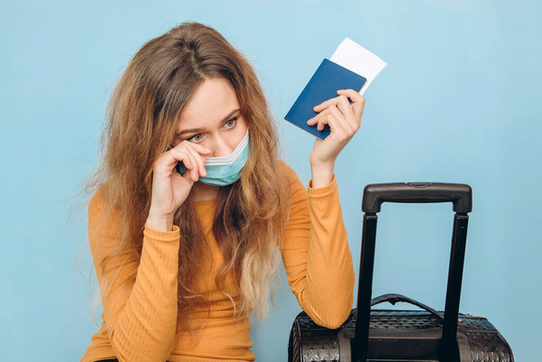 Chica turista en una máscara médica se sienta con el equipaje en sus manos sobre un fondo azul. Brote de coronavirus COVID-19. Concepto de viaje y corona. Un turista no puede irse debido a una pandemia
. - Foto, imagen