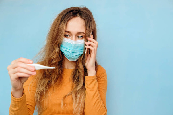 Close-up portrait of a girl in a medical mask on her face with suspected coronavirus and arm with outstretched thermometer. 2020 coronavirus epidemic. Covid-2019. Woman on a blue background. - Photo, Image