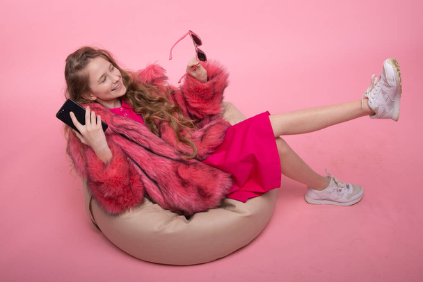 Mooi jong meisje zit in een fauteuil beige tas luistert naar muziek aan de telefoon. Modieuze kleding in roze kleuren. - Foto, afbeelding