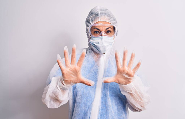 Middle age nurse woman wearing protection coronavirus equipment over white background afraid and terrified with fear expression stop gesture with hands, shouting in shock. Panic concept. - Photo, image