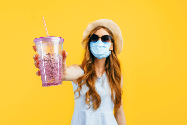 Happy girl in a summer hat and sunglasses, wears a medical protective mask against a viral infection, holds a cocktail in her hands on a yellow background. Quarantine, coronavirus, summer - Фото, зображення