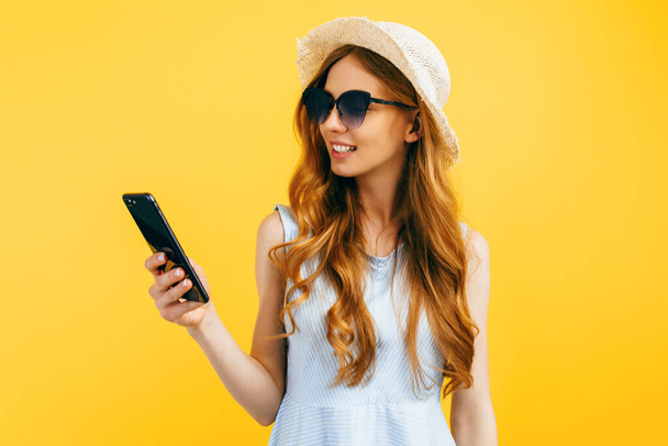 Happy beautiful girl in a summer hat, uses a mobile phone, standing on isolated blue background - Фото, изображение