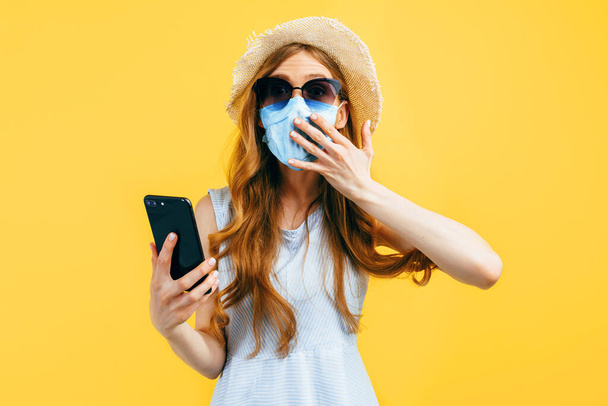 Shocked girl in a summer hat, wears a medical protective mask against a viral infection, uses a mobile phone on a yellow background. Quarantine, coronavirus, summer - Фото, изображение