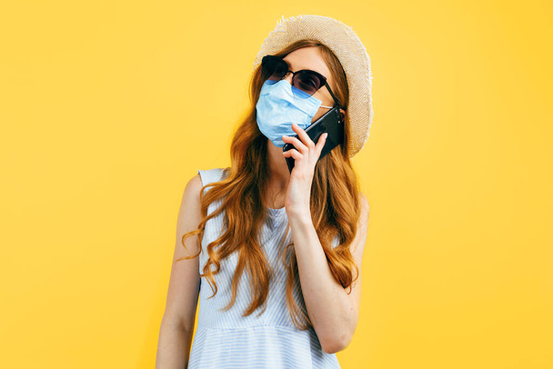 Shocked girl in a summer hat, wears a medical protective mask against a viral infection, talking on a cell phone on a yellow background. Quarantine, coronavirus, summer - Foto, Bild