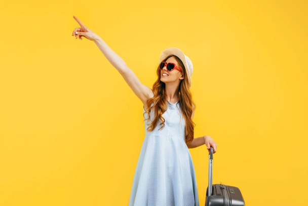 Attractive tourist girl in a summer hat standing with a suitcase isolated on a blue background. Travel, summer vacation concept - Foto, imagen