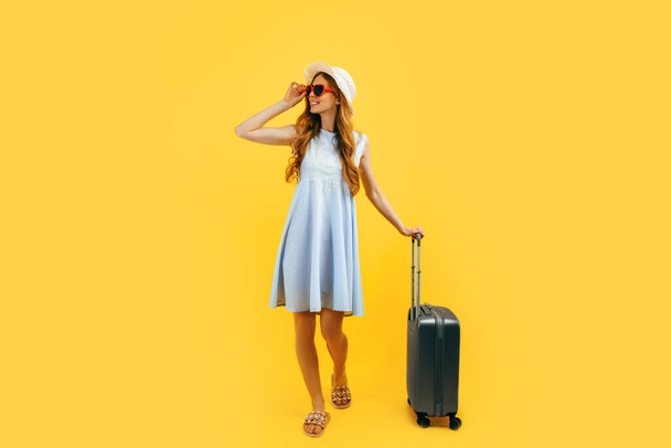 Attractive tourist girl in a summer hat standing with a suitcase isolated on a blue background. Travel, summer vacation concept - Foto, Bild
