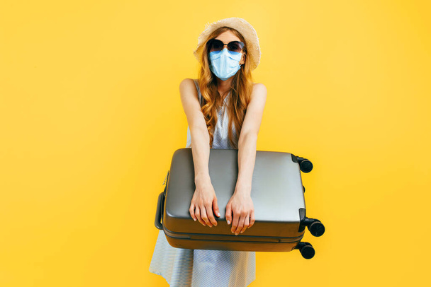 A young woman in a summer hat and a medical protective mask holds a suitcase in her hands on a blue background. The concept of quarantine, coronavirus, summer vacation - Фото, изображение