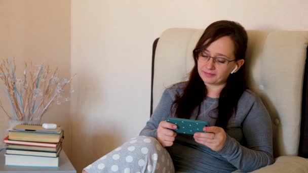 Усміхнена розслаблена жінка тримає смартфон, щоб дивитися відео сидіти на ліжку у вітальні. Щаслива дівчина в окулярах і навушниках дивиться на мобільний телефон
. - Кадри, відео