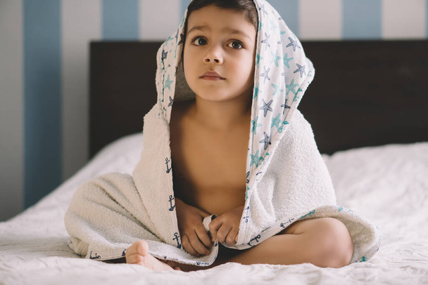 lindo niño, cubierto con toalla con capucha, sentado en la cama y mirando hacia otro lado
 - Foto, Imagen
