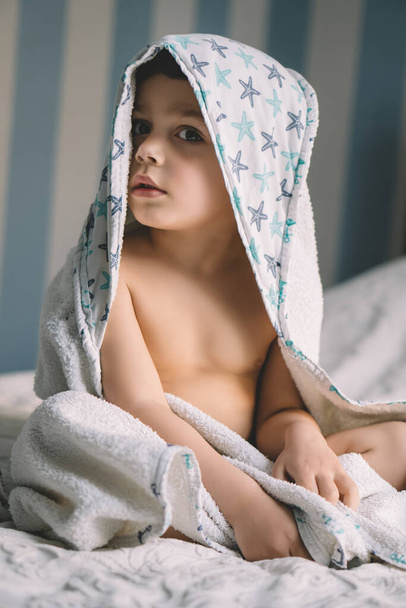 słodkie dziecko, zawinięte w zakapturzony ręcznik, siedzące na łóżku i patrzące w kamerę - Zdjęcie, obraz