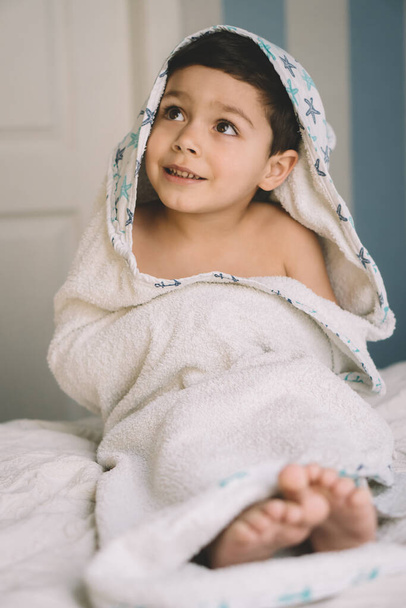 selectieve focus van gelukkig kind, verpakt in capuchon handdoek, weg te kijken terwijl zittend op bed - Foto, afbeelding
