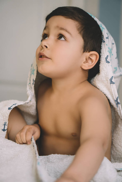 selective focus of adorable boy, wrapped in hooded towel, looking up while sitting on bed - Photo, Image