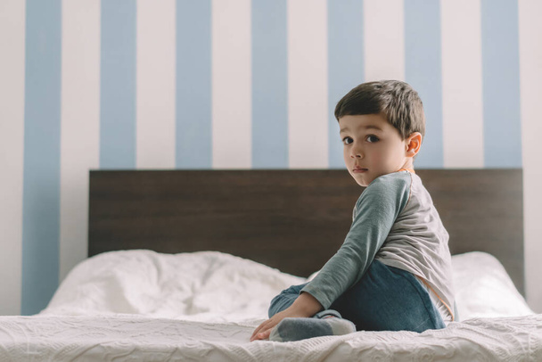 cute, serious boy looking at camera while sitting on bed at home - Фото, зображення