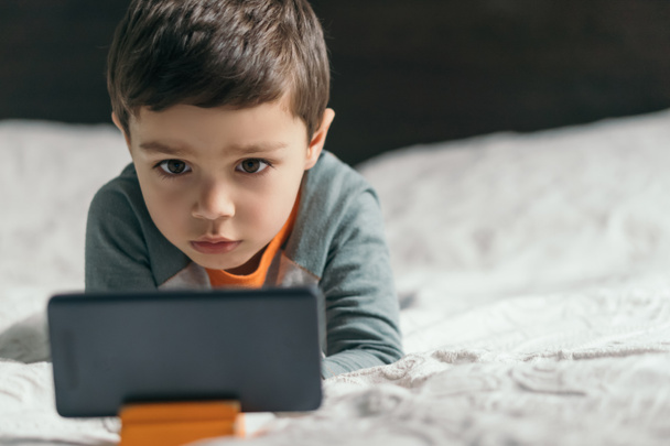 selective focus of adorable, focused boy watching online lesson on smartphone in bedroom - Foto, Imagem
