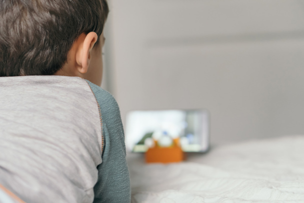 Rückansicht eines kleinen Jungen beim Webinar für Kinder auf dem Smartphone, während er im Bett liegt - Foto, Bild