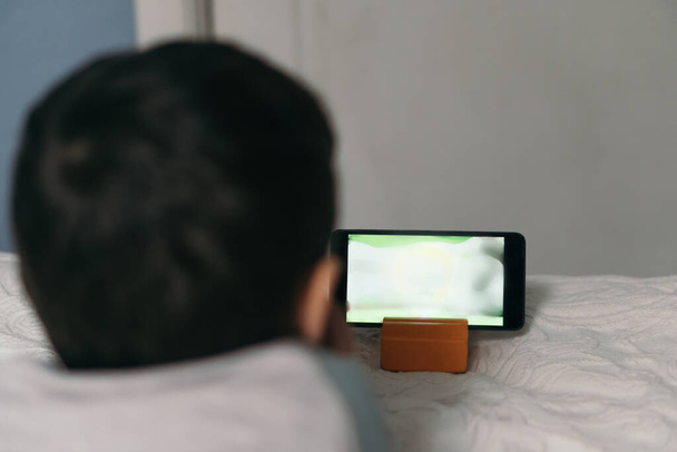 vista posterior del niño viendo dibujos animados educativos en el teléfono inteligente mientras está acostado en la cama
 - Foto, imagen