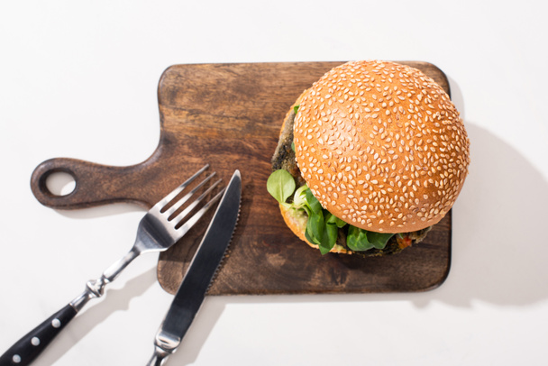 Draufsicht auf leckeren veganen Burger auf Holzschneidebrett mit Besteck auf weißem Hintergrund - Foto, Bild