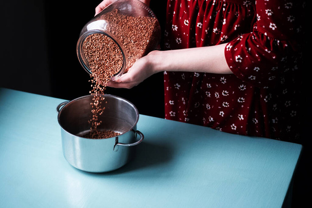 Process how hands of a young woman in a red dress pour buckwheat groats from a large glass jar into a pan on a blue table. Vew from above. Pandemic Deficit - 写真・画像