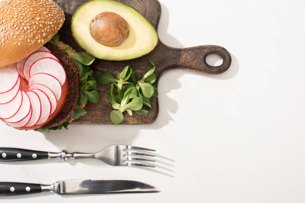 top view of delicious vegan burger with radish, avocado and greens on wooden cutting board near cutlery on white background - Photo, Image