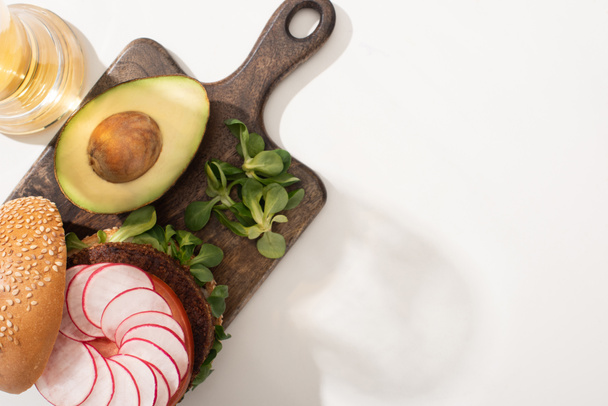 top view of delicious vegan burger with radish, avocado and greens on wooden cutting board on white background - Photo, Image
