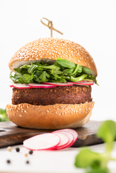 selective focus of delicious vegan burger with radish and arugula on wooden board with black pepper on white background - Zdjęcie, obraz