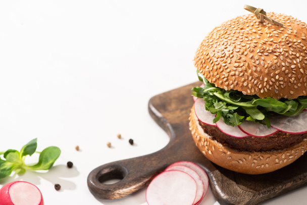 selective focus of  delicious vegan burger with radish and arugula on wooden board with black pepper on white background - Photo, Image