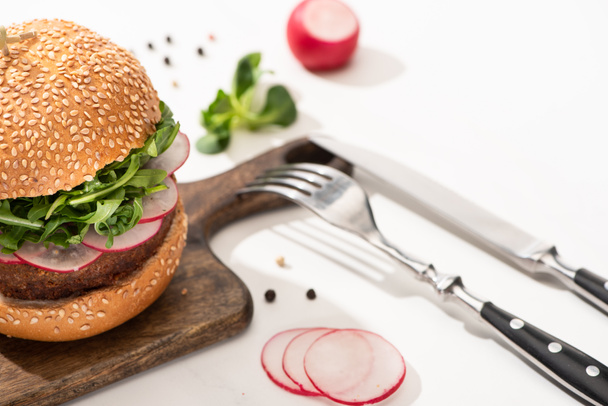 selective focus of delicious vegan burger with radish and arugula on wooden board with black pepper near fork and knife on white background - Photo, Image