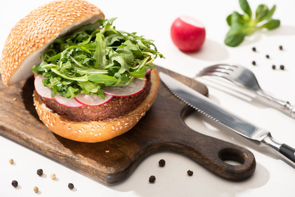 selective focus of delicious vegan burger with radish and arugula on wooden board with black pepper near fork and knife on white background - Foto, Imagem