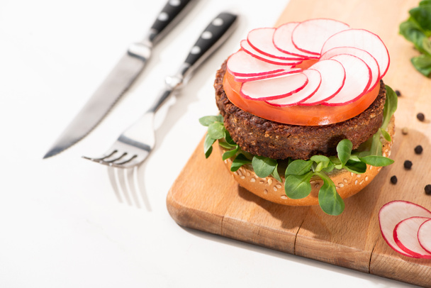selective focus of delicious vegan burger with radish, tomato and microgreens on wooden boar with black pepper near fork and knife on white background - 写真・画像