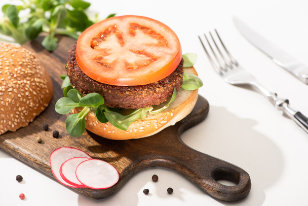selektivní zaměření lahodného veganského burgeru s ředkvičkami, rajčaty a mikrozelení na dřevěném kanci s černým pepřem u vidličky a nožem na bílém pozadí - Fotografie, Obrázek